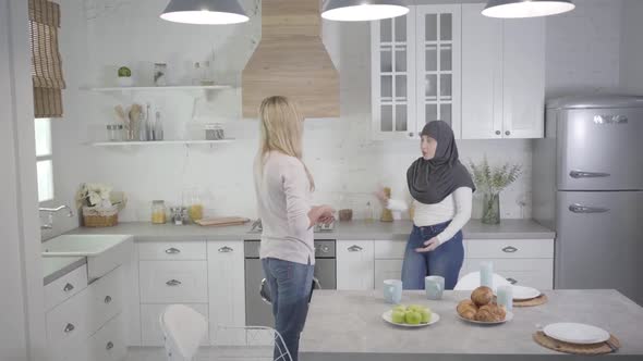 Muslim Woman in Hijab Arguing with Blond Caucasian Friend at Home. Two Women Quarrelling Indoors alt