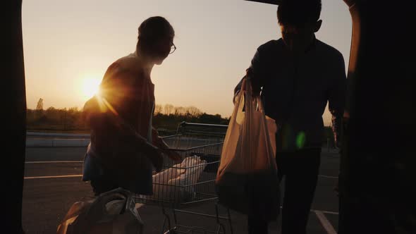 A Man and a Woman Are Unloading Purchases From a Trolley in the Trunk of a Car. Buying Food alt