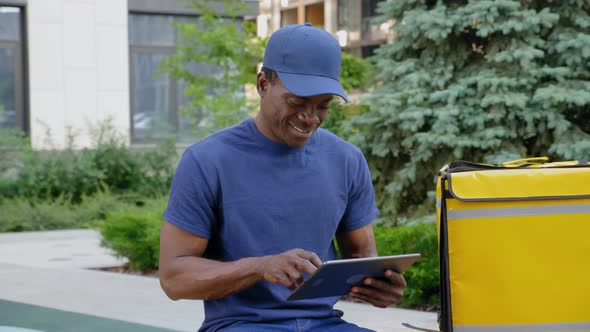 Smiling Afroamerican Man Courier Food Delivery Sitting on Bench Uses Tablet alt