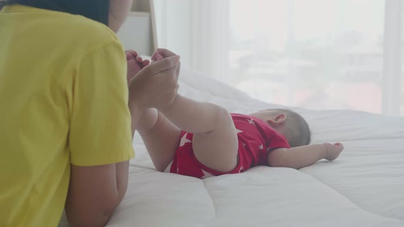 Young asian mother kiss foot of little baby girl with tender on bed in the bedroom. alt