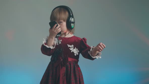 Little Girl in Big Headphones and Vintage Dress is Listening to Music on Phone