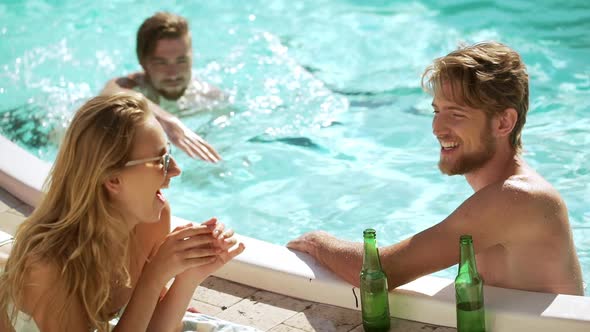 Two Caucasian Muscle Males in Pool Talking Laughing Smiling with Blonde Female Lying on Lounge alt