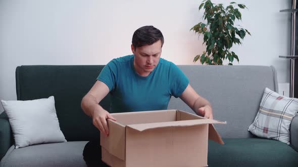 A man opens a cardboard box while sitting on a home office couch.