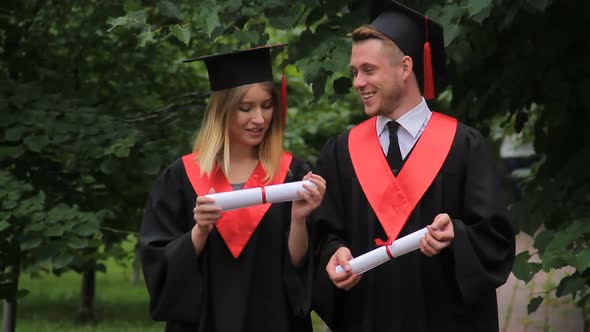 Woman and Man in Academic Dresses Holding Diplomas Talking and Walking in Park alt