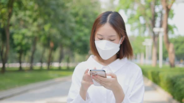 A woman wearing a protective mask using a smartphone as she walks on the pavement.