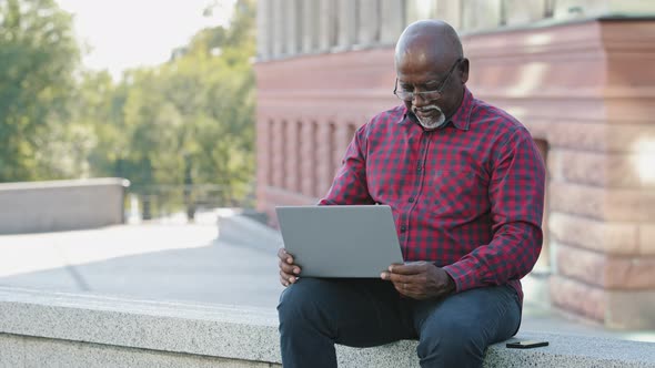 African American Mature Man In Shirt Sitting Outdoors Using Laptop Communicating on Social Networks alt