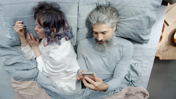 Mature Man Husband Using Smartphone Texting While Young Woman Sleeping in Bed alt