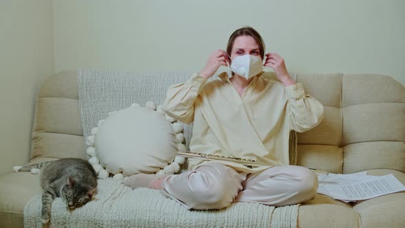 Woman musician with flute in medical face mask playing at home on sofa in living room