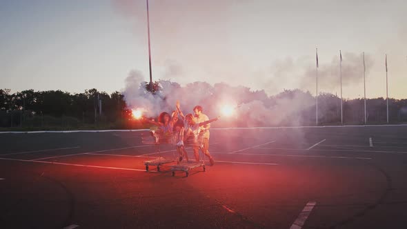 Young Friends Riding on Shopping Trolleys at Deserted Parking Lot of Hypermarket Holding Burning alt