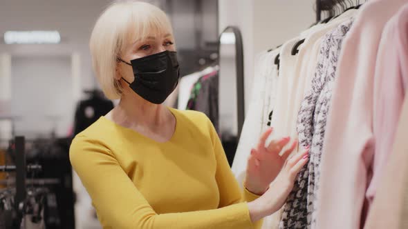 Elderly Woman Buyer in Protective Mask Chooses Clothes in Boutique Looking for New Outfit Happy alt