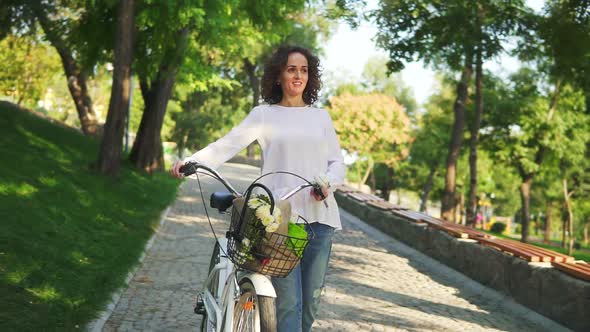 Smiling Attractive Woman in a White Tshirt and Blue Jeans Walking Holding Her City Bicycle's alt