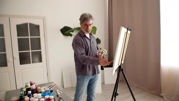 Man Holds Palette in Hand and Draws with Brush on Canvas Standing in Living Room There are Many Cans alt