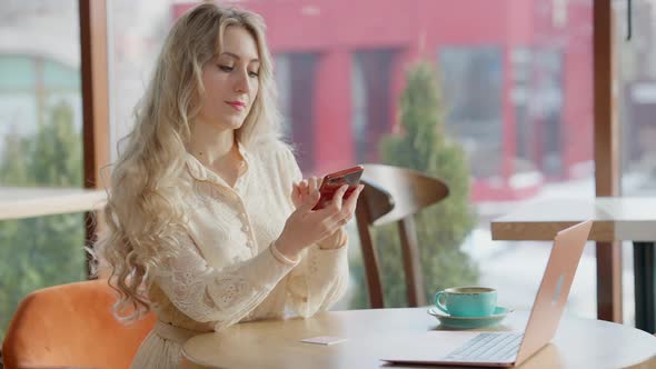 Elegant Slim Beautiful Woman Sitting in Cafe Surfing Internet on Smartphone alt