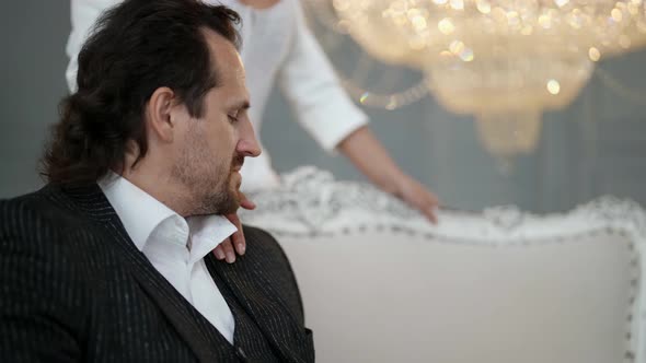 Bride is Stroking Shoulder of Her Beloved Groom Sitting on Couch Solemn Wedding Day alt