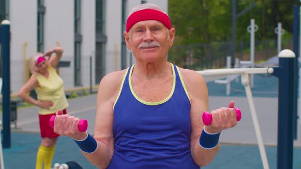 Senior Man Grandfather Doing Training Weightlifting Exercising with Dumbbells on Playground in Park alt