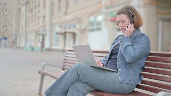 Old Woman Talking on Phone and Using Laptop While Sitting Outdoor on Bench alt