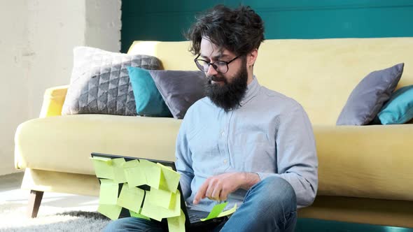 Man Working at the Computer with a Lot of Reminder Notes Sitting Near Sofa alt