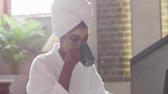 African american attractive woman with applied face mask drinking coffee and using laptop at home alt