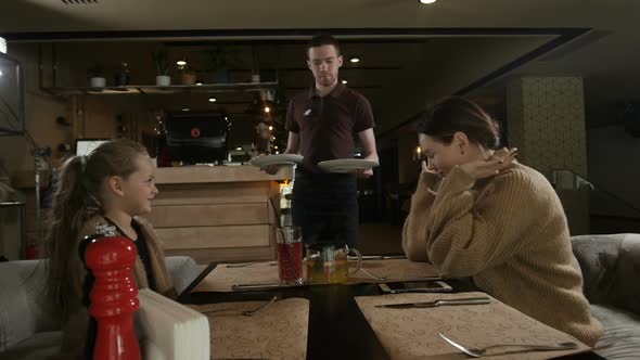 a Man Waiter Brings Food to His Mother and Daughter Sitting in a Cafe alt
