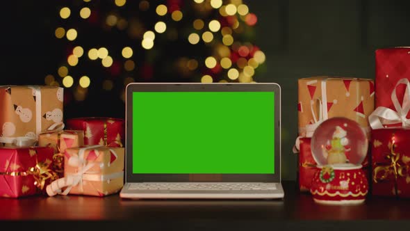 Laptop with Green Screen on Table with Christmas Decorations Against Christmas Tree