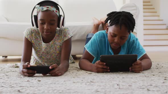 African american brother and sister using electronic devices lying on the floor at home alt
