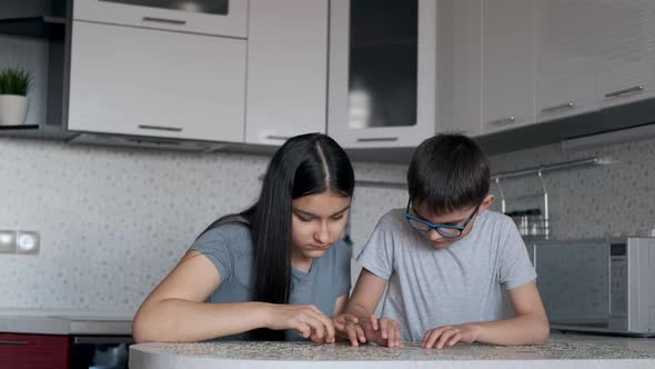 Cheerful Boy and Girl Will Put Together a Puzzle While Sitting at a Table at Home in the Kitchen alt