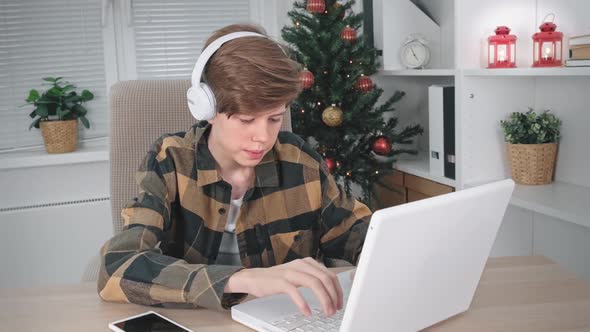 A Teenage Boy with Headphones is Typing on a Laptop Studying Information on the Internet Sitting at