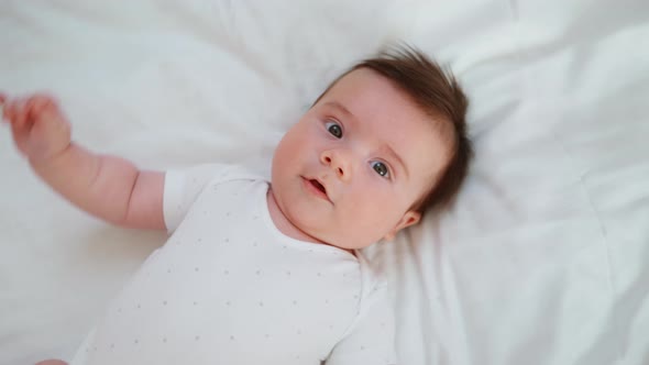 A Newborn Baby Lies on His Back on a White Bed and Looks at the Camera