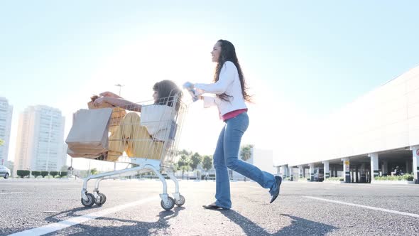 Young woman pushing the grocery cart, while her friend is sitting inside during sunset.