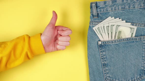 Female hand showing thumbs up against a background of many dollars in a jeans pocket