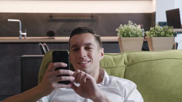 Happy Man Falls on the Couch After Work and Uses Smartphone While Enjoying His Vacation at Home alt