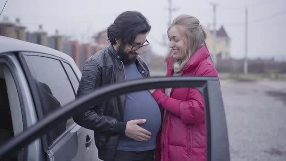 Smiling Caucasian Blond Woman Caressing Her Husband's Pregnant Belly and Imitating Listening Baby alt