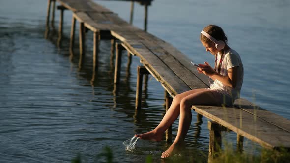 A little girl wearing headphones with a smartphone sits on a wooden pier on the lake 