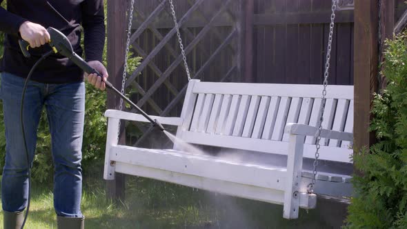 Caucasian Man Washes Dirt Off a Wooden Bench Swing Using a High Pressure Washer alt