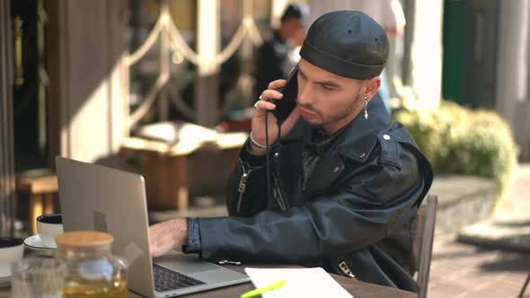 Busy Caucasian Gay Man Talking on Phone Messaging Online on Laptop Sitting in Cafe Outdoors alt