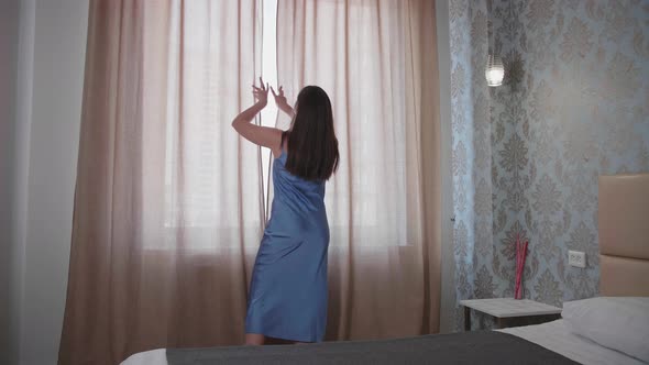 Young Woman Approaches the Window in the Bedroom Pushes Beige Curtains with Her Hands and Enjoys