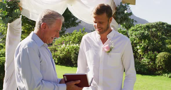 Smiling caucasian senior male wedding officiant holding book and groom standing in outdoor altar alt