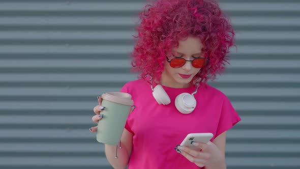 A Pink Curly Teenager is Using a Smartphone Outdoors alt