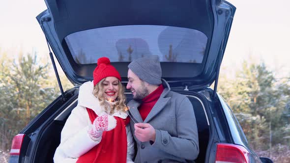 Young Caucasian Couple Sitting in Opened Car with Sparklers Snowy Forest alt