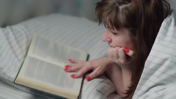 Pretty Brunette Woman Lies on the Bed and Reads a Book Covered with a Blanket at Night Spends Time