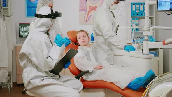 Pediatric Dentist in Ppe Suit Talking with Kid Patient Writing on Clipboard alt