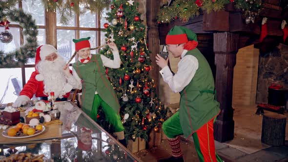 Santa with funny elves at Christmas. Elf is taking photo of Santa and girl elf on Christmas room alt