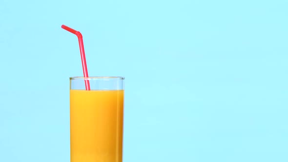 Glass of Orange Juice with Drinking Tube Rotating on Blue Background alt