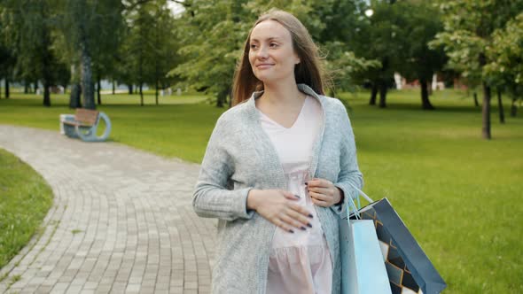 Happy Pregnant Girl Walking in City Park Holding Shopping Bags Smiling on Summer Day alt