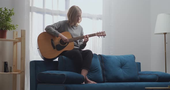 A Young Girl in a Gray Sweatshirt and Black Pants Composes New Music on an Acoustic Guitar alt
