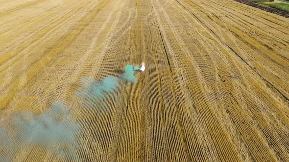 Blond woman in white dress running in a field with green smoke grenade alt