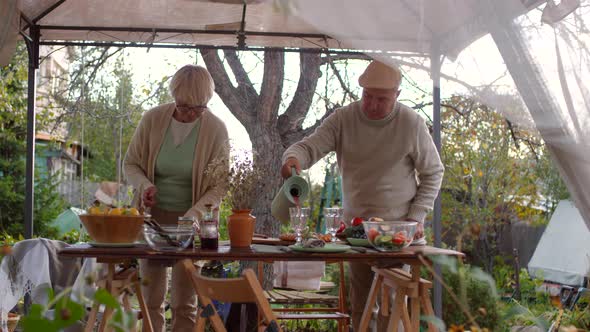 Elderly Caucasian Couple Setting up Lunch in Garden Gazebo alt