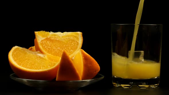 Juice Is Poured Into A Glass With Ice, Several Sliced Oranges In A Plate On A Black Background