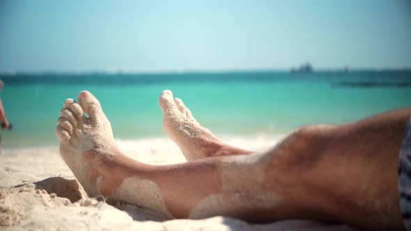 Man Lying On Beach And Moving Legs On Holiday Vacation. Legs Man On Beach Sun Lounger Sandy Shore. alt
