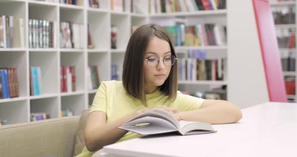 Young Beautiful European Female Student Wearing Glasses is Reading Book in College Library
